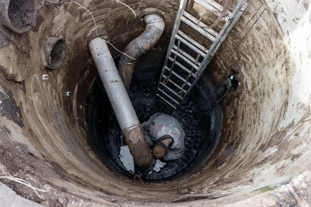 Person inside a deep cesspool looking up with a ladder and pipe.